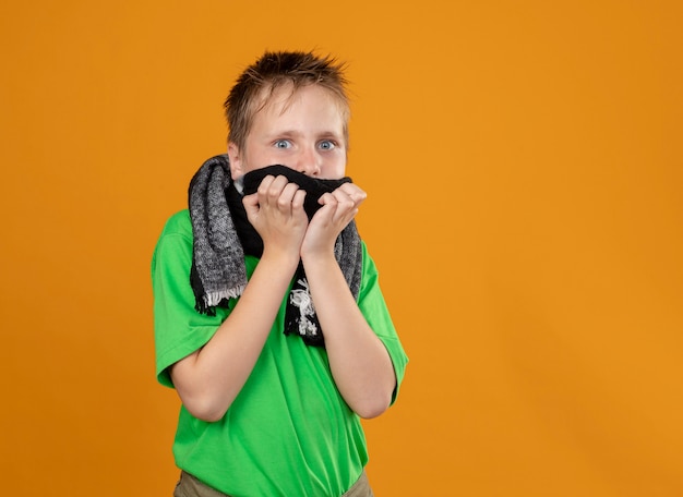 Petit garçon malade en t-shirt vert et écharpe chaude autour du cou et de la bouche en panique debout sur un mur orange