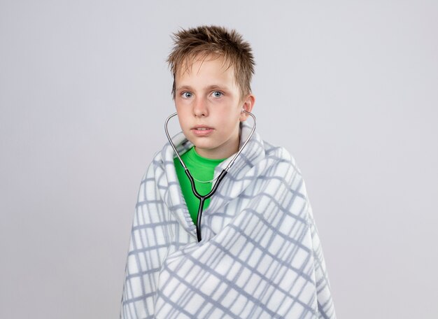 Petit garçon malade en t-shirt vert enveloppé dans une couverture se sentant mal avec stéthoscope à debout sur un mur blanc