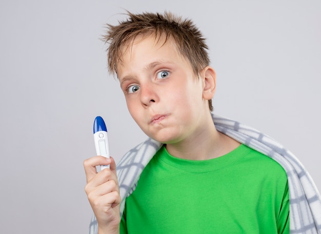 Petit garçon malade en t-shirt vert enveloppé dans une couverture se sentant mal tenant un thermomètre à l'inquiétude et étonné debout sur un mur blanc