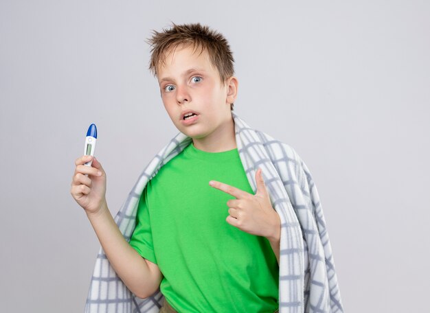 Petit garçon malade en t-shirt vert enveloppé dans une couverture se sentant mal tenant un thermomètre pointant avec le doigt sur le fait d'être inquiet debout sur un mur blanc