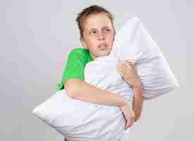Photo gratuite petit garçon malade en t-shirt vert se sentant mal à l'aise étreignant oreiller à côté impitoyable et malade debout sur mur blanc