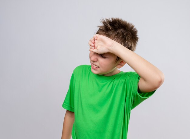 Petit garçon malade en t-shirt vert se sentir mal au front de toucher souffrant de froid debout sur un mur blanc