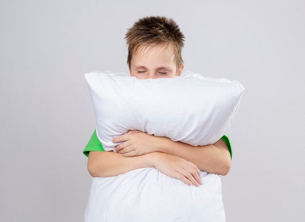 Petit garçon malade en t-shirt vert se sentir mal en serrant l'oreiller avec les yeux fermés debout sur un mur blanc