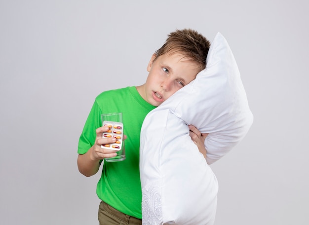 Petit garçon malade en t-shirt vert se sentir mal tenant un oreiller et un verre d'eau appuyé sur la tête sur l'oreiller debout sur un mur blanc
