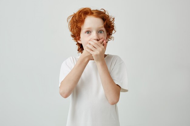 Petit garçon mignon au gingembre aux yeux verts dans la bouche de vêtements de t-shirt blanc avec les mains, ayant peur de regarder un film d'horreur avec des amis.