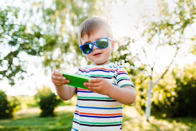 Un petit garçon mignon tape un message sur son téléphone avec des lunettes de soleil lumineuses.