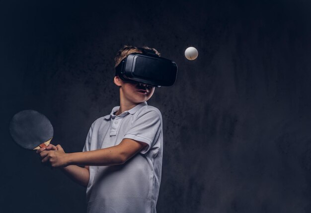 Petit garçon roux vêtu d'un t-shirt blanc jouant au ping-pong avec des lunettes de réalité virtuelle. Isolé sur un fond texturé sombre.