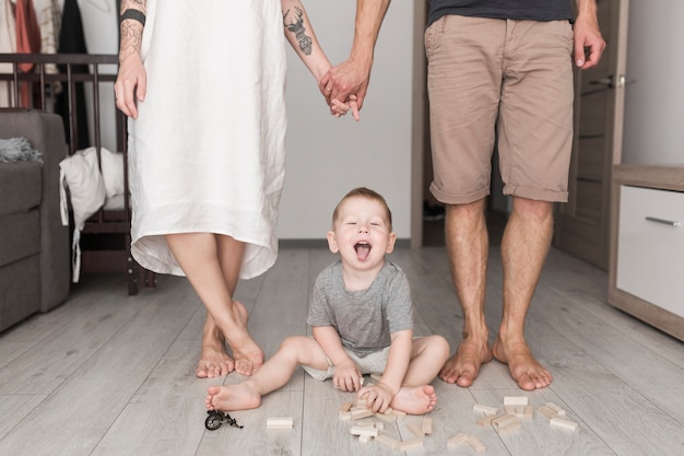 Petit garçon s&#39;amuse avec des blocs de bois avec ses parents debout derrière lui, se tenant la main