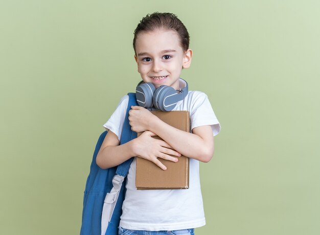 Petit garçon souriant portant un sac à dos et des écouteurs autour du cou tenant et étreignant un livre
