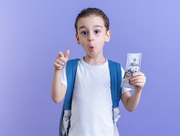 Photo gratuite petit garçon surpris portant un sac à dos tenant un dollar regardant et pointant vers la caméra isolée sur un mur violet