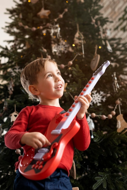 Photo gratuite petit garçon tenant une guitare à côté de l'arbre