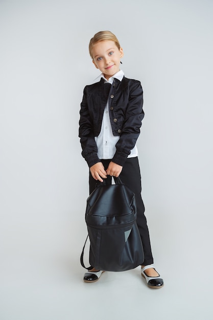 Petit modèle féminin posant en uniforme scolaire avec sac à dos sur mur blanc