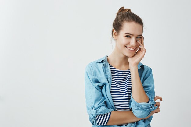 Une petite amie attrayante ne peut pas arrêter de regarder son petit ami. Mince femme caucasienne avec coiffure chignon touchant doucement la joue avec la paume et souriant joyeusement tout en se tenant sur le mur gris.