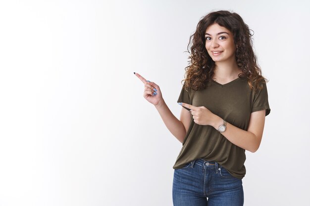 Petite amie curieuse mignonne demandant ce qu'il y a vers la gauche intéressée indiquer l'index souriant intrigué excité debout fond blanc voulant regarder de plus près le produit