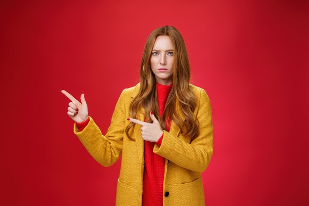 Petite amie mécontente, offensée et sombre, posant une question, faisant la moue et fronçant les sourcils à cause de l'insulte pointant vers le coin supérieur gauche déçue et contrariée posant insatisfaite sur fond rouge.