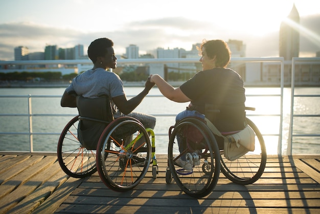 Photo gratuite petite amie et petit ami qui passent la journée ensemble. homme afro-américain et femme de race blanche en fauteuil roulant, main dans la main, regardant le coucher du soleil. amour, affection, concept de bonheur