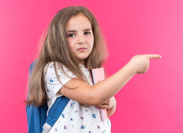 Photo gratuite petite belle fille aux cheveux longs avec sac à dos tenant un cahier pointant avec l'index sur le côté regardant à l'avant avec un visage sérieux debout sur un mur rose