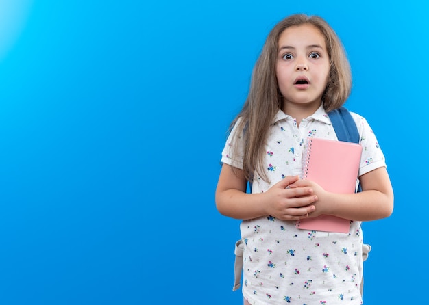 Petite belle fille aux cheveux longs avec sac à dos tenant un cahier regardant devant étonné et surpris debout sur le mur bleu