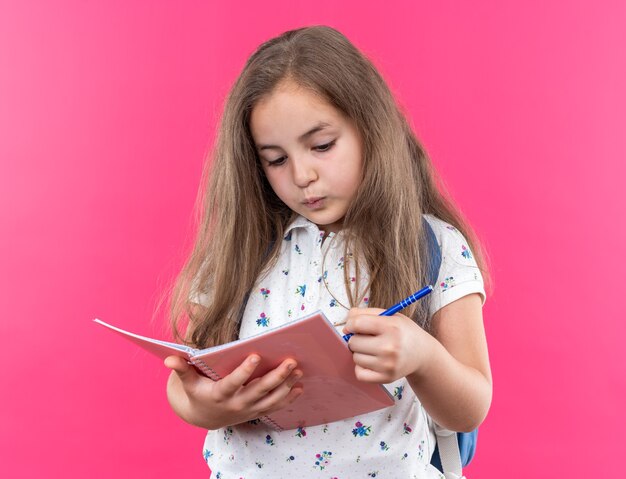 Petite belle fille aux cheveux longs avec sac à dos tenant un cahier et un stylo à l'air confiant debout sur le rose