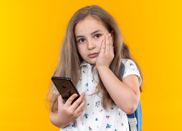 Petite belle fille aux cheveux longs avec sac à dos tenant un smartphone inquiet debout sur orange