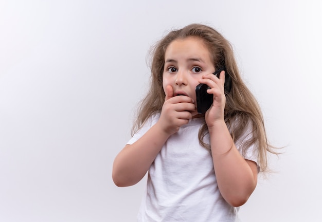 Petite écolière effrayée portant un t-shirt blanc parle sur la bouche couverte de téléphone sur fond blanc isolé