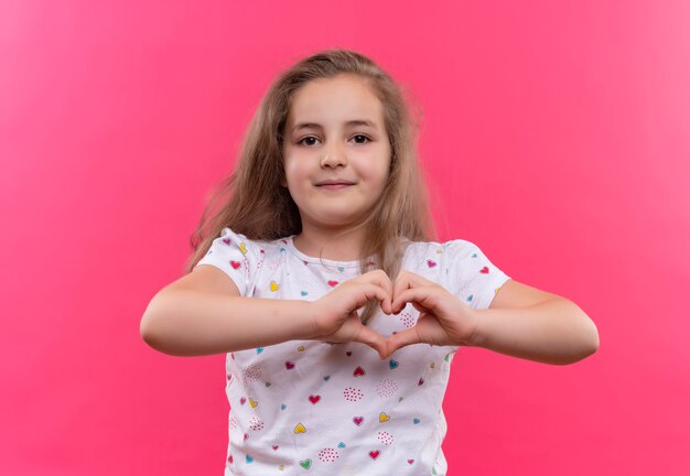 Petite écolière portant un t-shirt blanc montrant le geste du cœur sur un mur rose isolé