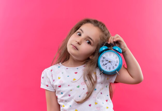 Petite écolière portant un t-shirt blanc tenant un réveil et écouter sur un mur rose isolé