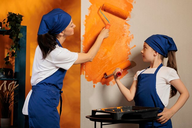 Petite famille utilisant une brosse à rouleau pour peindre, peignant les murs de la pièce avec une couleur orange. Adulte et petite fille rénovant l'intérieur de l'appartement avec un pinceau et des outils de bricolage, amélioration de la décoration des travaux ménagers.