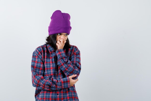 Petite femme en chemise à carreaux et bonnet tenant la main sur la bouche à la perplexité