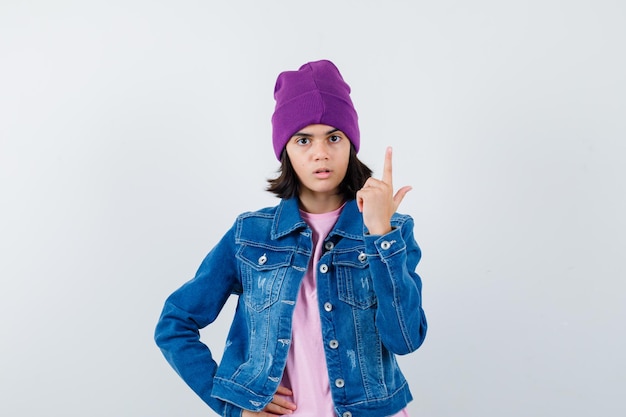 Petite femme pointant vers le haut dans une veste en jean et un bonnet à la perplexité