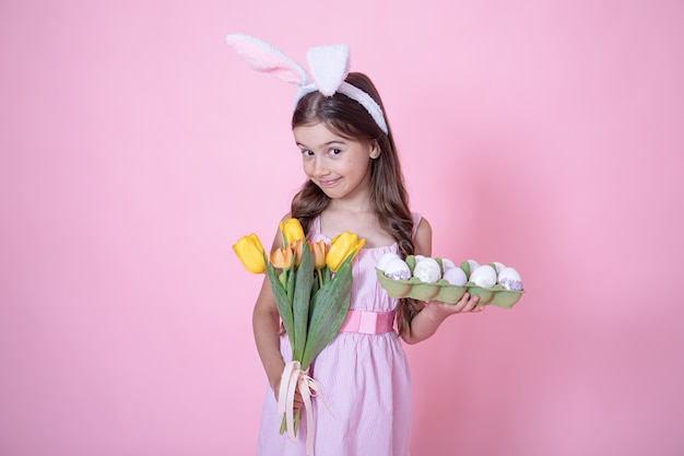 Petite fille aux oreilles de lapin de Pâques tient un bouquet de tulipes et un plateau d'oeufs dans ses mains sur rose