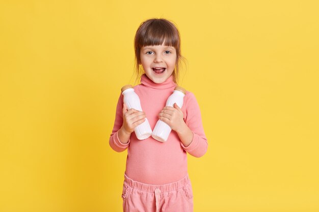 Petite fille de beauté dans des vêtements roses aux cheveux bruns tenant deux bouteilles de lait sur jaune