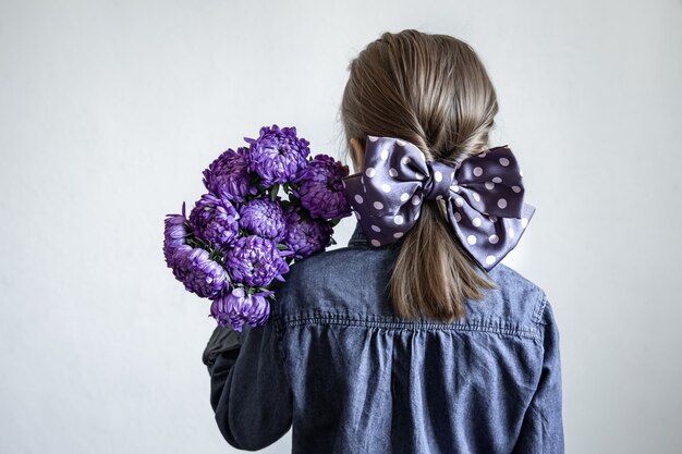 Petite fille avec un bel arc sur ses cheveux tient un bouquet de chrysanthèmes bleus, vue arrière.