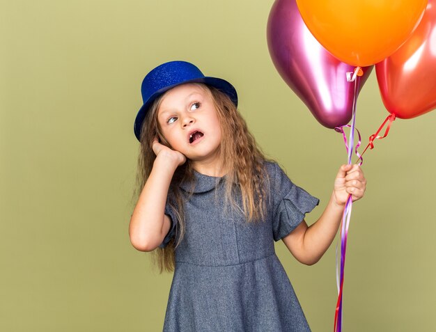 Petite fille blonde anxieuse avec un chapeau de fête bleu tenant des ballons à l'hélium et regardant le côté isolé sur un mur vert olive avec espace pour copie