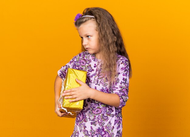 petite fille blonde confiante tenant une boîte-cadeau et regardant le côté isolé sur un mur orange avec espace de copie