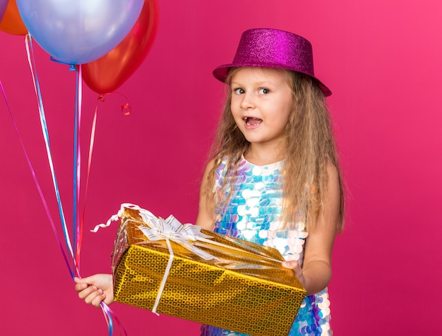 petite fille blonde excitée avec un chapeau de fête violet tenant des ballons à l'hélium et une boîte-cadeau isolée sur un mur rose avec un espace de copie