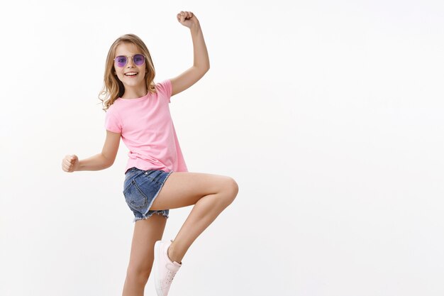 Petite fille blonde gaie et charismatique dans des lunettes de soleil d'été, t-shirt rose sautant, soulevant la jambe posant joyeusement, dansant en s'amusant, levant les mains amusées, debout mur blanc heureux