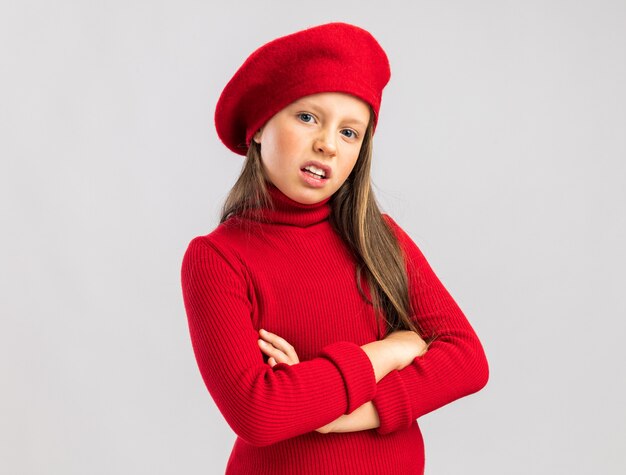 Petite fille blonde inquiète portant un béret rouge gardant les bras croisés regardant devant isolé sur mur blanc avec espace de copie