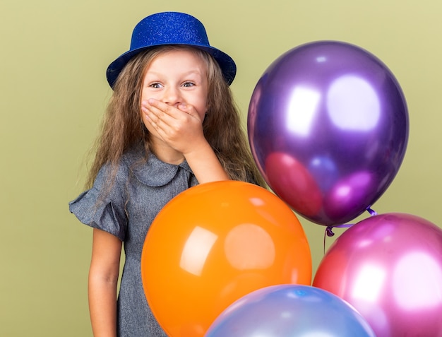 petite fille blonde surprise avec un chapeau de fête bleu mettant la main sur la bouche et tenant des ballons à l'hélium isolés sur un mur vert olive avec espace de copie