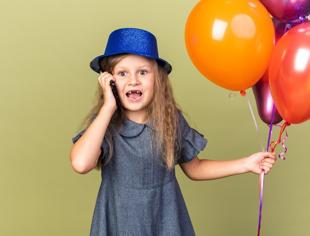 petite fille blonde surprise avec un chapeau de fête bleu tenant des ballons à l'hélium et parlant au téléphone isolé sur un mur vert olive avec espace de copie