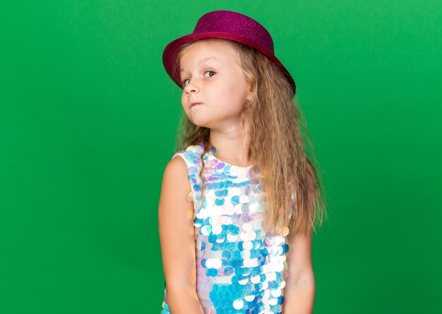 petite fille blonde surprise avec un chapeau de fête violet regardant le côté isolé sur un mur vert avec espace pour copie