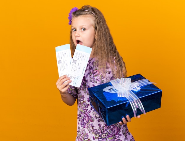 petite fille blonde surprise tenant une boîte-cadeau et des billets d'avion isolés sur un mur orange avec espace de copie