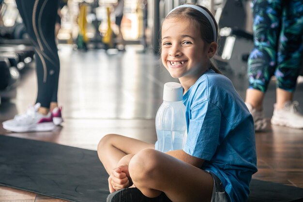 Une petite fille avec une bouteille d'eau est assise sur un tapis dans la salle de gym