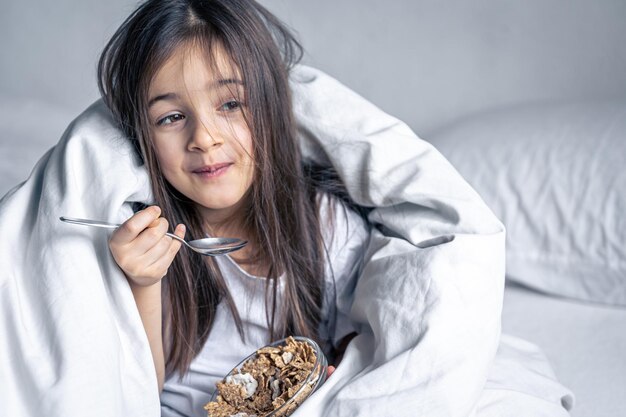 Petite fille brune mignonne dans un lit blanc a des céréales pour le petit déjeuner