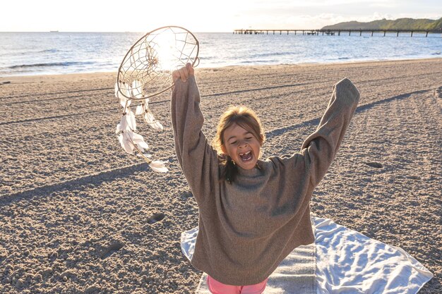 Photo gratuite petite fille avec un capteur de rêves au bord de la mer au coucher du soleil