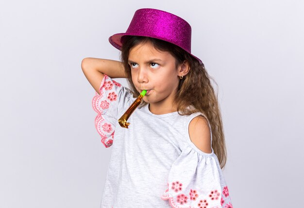 petite fille caucasienne agacée avec un chapeau de fête violet soufflant un sifflet de fête et levant isolé sur un mur blanc avec espace de copie