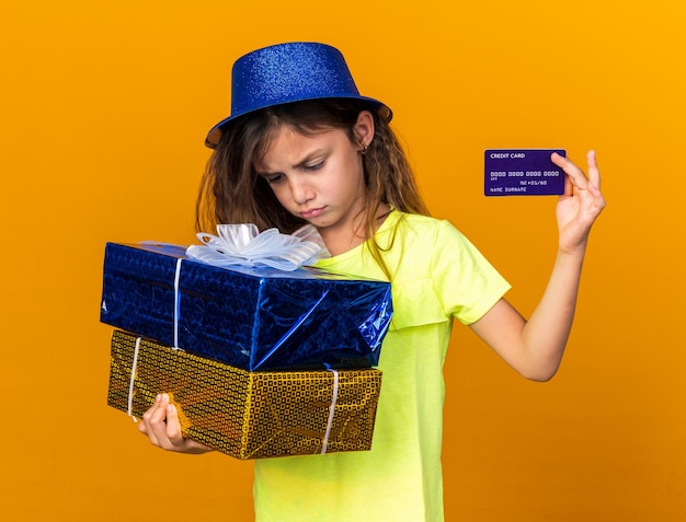 petite fille caucasienne déçue avec un chapeau de fête bleu tenant des coffrets cadeaux et une carte de crédit isolée sur un mur orange avec espace de copie