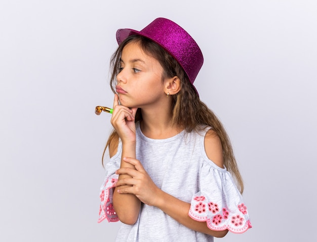petite fille caucasienne réfléchie avec un chapeau de fête violet mettant la main sur le menton tenant un sifflet de fête et regardant le côté isolé sur un mur blanc avec un espace de copie