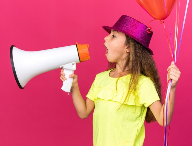 petite fille caucasienne surprise avec un chapeau de fête violet tenant des ballons à l'hélium et parlant dans un haut-parleur regardant le côté isolé sur un mur rose avec un espace de copie