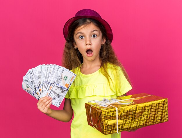 petite fille caucasienne surprise avec un chapeau de fête violet tenant une boîte-cadeau et de l'argent isolé sur un mur rose avec un espace de copie